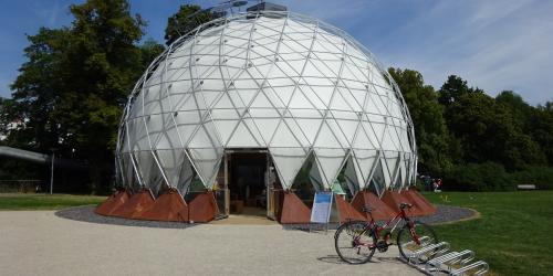 Kuppelartiges helles temporäres Bauwerk mit einem sichtbaren Eingang steht vor einer Wiese. Rechts davor ein Fahrradständer mit einem Fahrrad