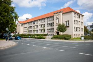 Blick auf eine Kreuzung. Auf der Linkesn Seite stehen Uatos an einer Ampel. Im Hintergund ist ein großes Gebäude mit einem roten Spizdach zu sehen.