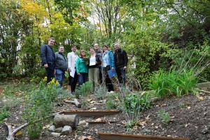 Acht Personen stehen in einem Garten und lächeln in die Kamera. Eine Person hat eine Urkunde und einen Blumenstrauß in der Hand.