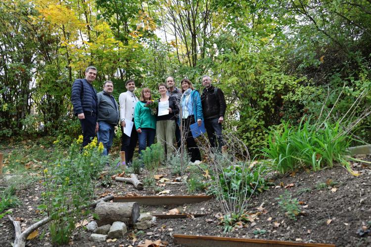 Acht Personen stehen in einem Garten und lächeln in die Kamera. Eine Person hat eine Urkunde und einen Blumenstrauß in der Hand.