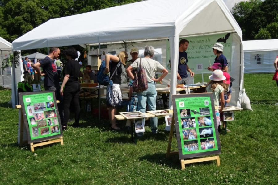 weißer Pavillon mit interessierten Menschen darunter und 2 Aufstellern davor