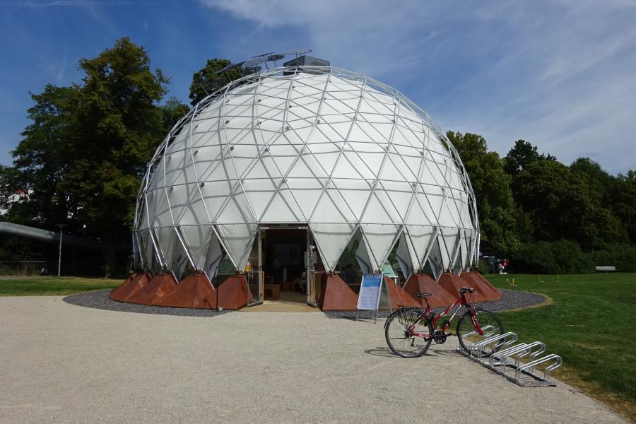 Kuppelartiges helles temporäres Bauwerk mit einem sichtbaren Eingang steht vor einer Wiese. Rechts davor ein Fahrradständer mit einem Fahrrad