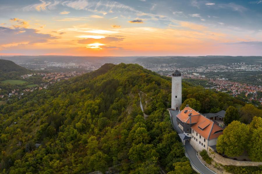 Blick aus der Vogelperspektive auf einen historischen Bergfried, der sich auf einem Berg befindet. Im dahinter gelegenen Tal erstreckt sich die Stadt Jena im Abendlicht.